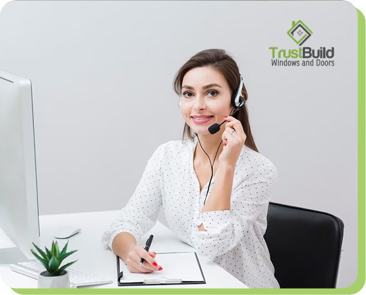 A smiling customer service representative wearing a headset sits at a white desk with a computer and notepad. The TrustBuild Windows and Doors logo is visible in the top right corner.