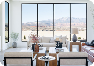 A bright, modern living room featuring four large floor-to-ceiling windows with thin black frames that offer a panoramic view of a desert mountain landscape.