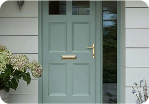 A traditional sage green front door featuring six recessed panels, two small glass windows at the top, and gold hardware including a letterbox and lever handle. It is installed in a white-sided house next to a flowering hydrangea bush.