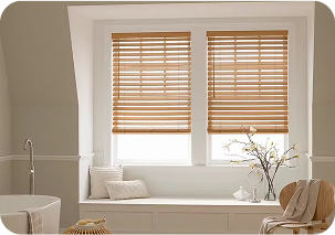 Light wood-toned Venetian blinds with horizontal slats installed on two adjacent windows in a modern bathroom, featuring a white window seat with decorative pillows and a bathtub in the foreground.