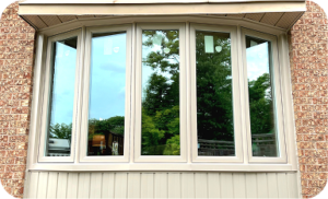 An exterior view of a newly installed five-panel bay window with cream-colored frames set into a red brick wall. The glass reflects the surrounding green trees and sky.