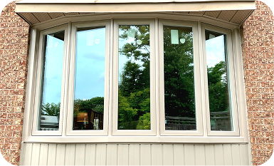An exterior view of a newly installed five-panel bay window with cream-colored frames set into a red brick wall. The glass reflects the surrounding green trees and sky.