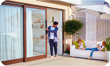 A man opening large wood-framed glass sliding patio doors that lead from a bright interior to a sunlit outdoor deck where two young children are sitting on a white sofa.