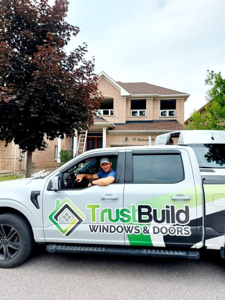 A TrustBuild Windows and Doors pickup truck is parked in front of a brick house. A technician smiles from the driver’s seat while window installation work is visible on the home's second floor.