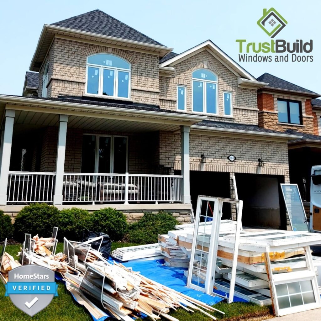 A large two-story stone house sits behind a pile of old window frames and debris on a blue tarp. New white-framed windows with arched tops are already installed on the home's second floor.