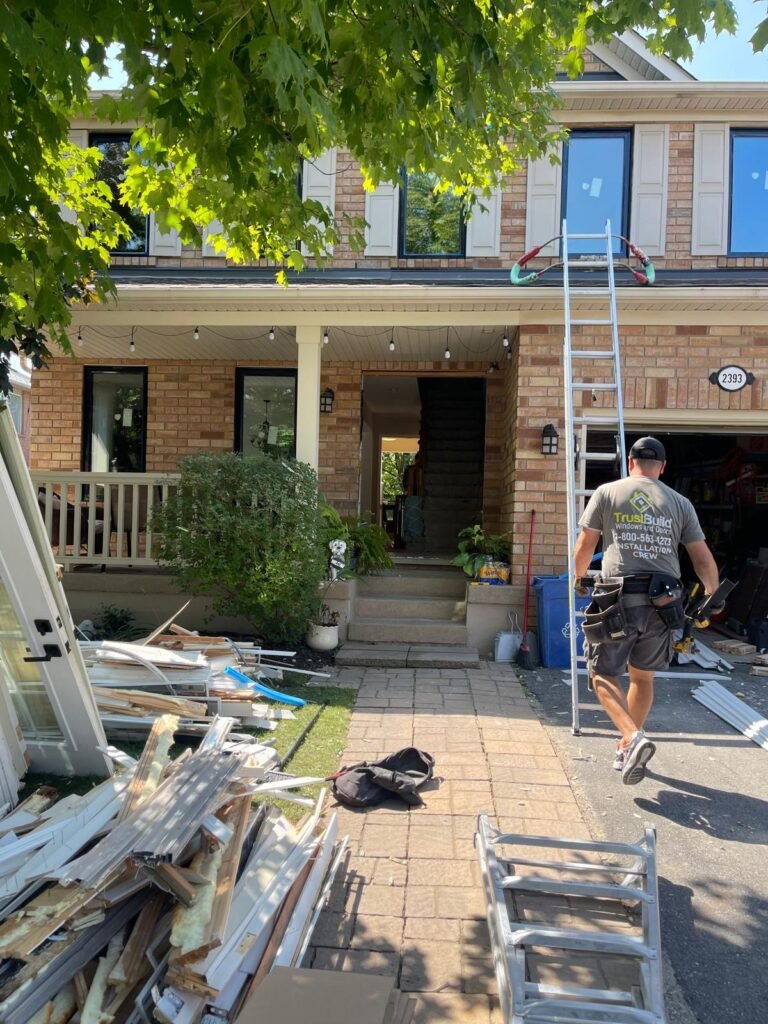 A worker with a tool belt walks toward a brick house during a window installation. Debris is piled on the lawn near a ladder leaning against the second story under a leafy green tree.