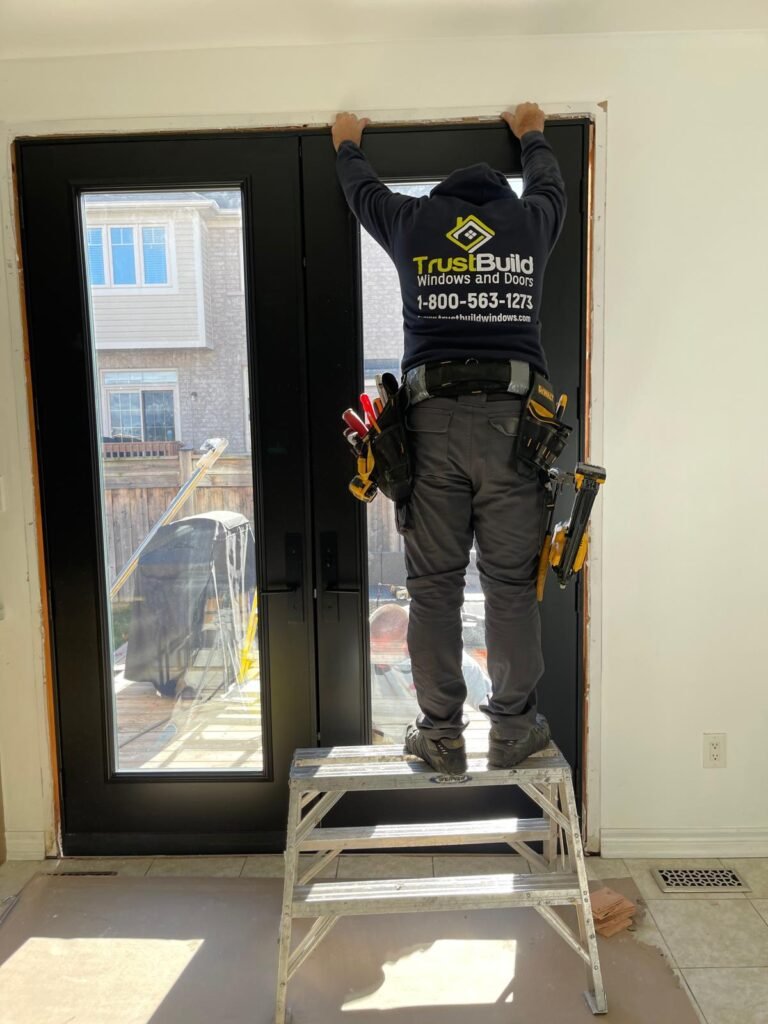 An installer in a navy TrustBuild hoodie stands on a stepladder to secure the top of a new black double-patio door. The interior installation overlooks a sunny backyard and deck.