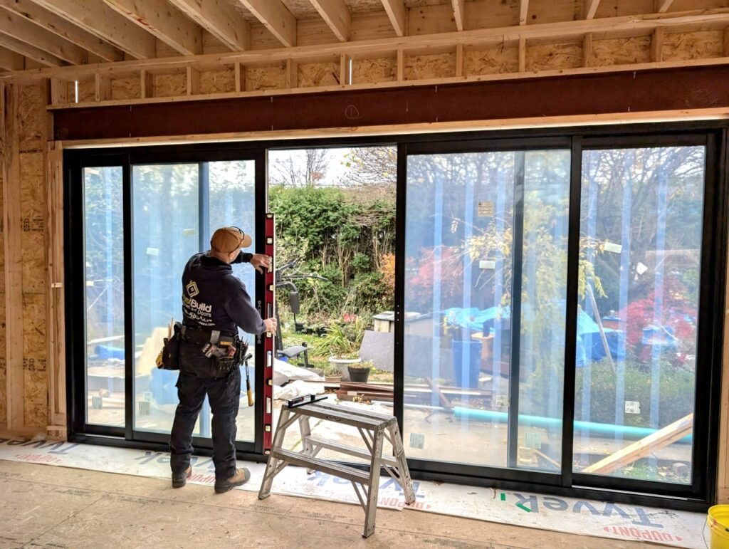 A TrustBuild Windows and Doors contractor uses a level to install large black sliding glass doors in a home under construction, with a view of a backyard garden through the glass panes.