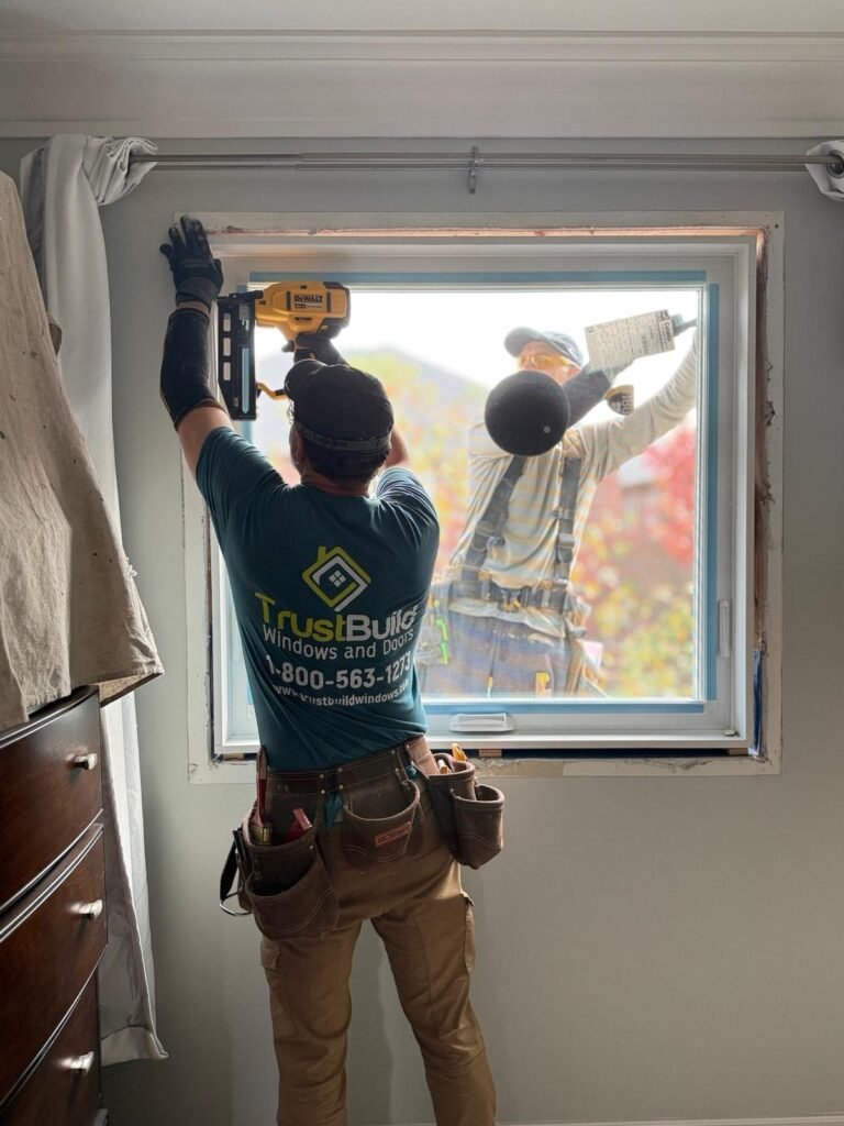 TrustBuild Windows and Doors contractors install a new window. One worker inside uses a nail gun on the frame, while another stands outside, viewed through the glass in a bedroom.