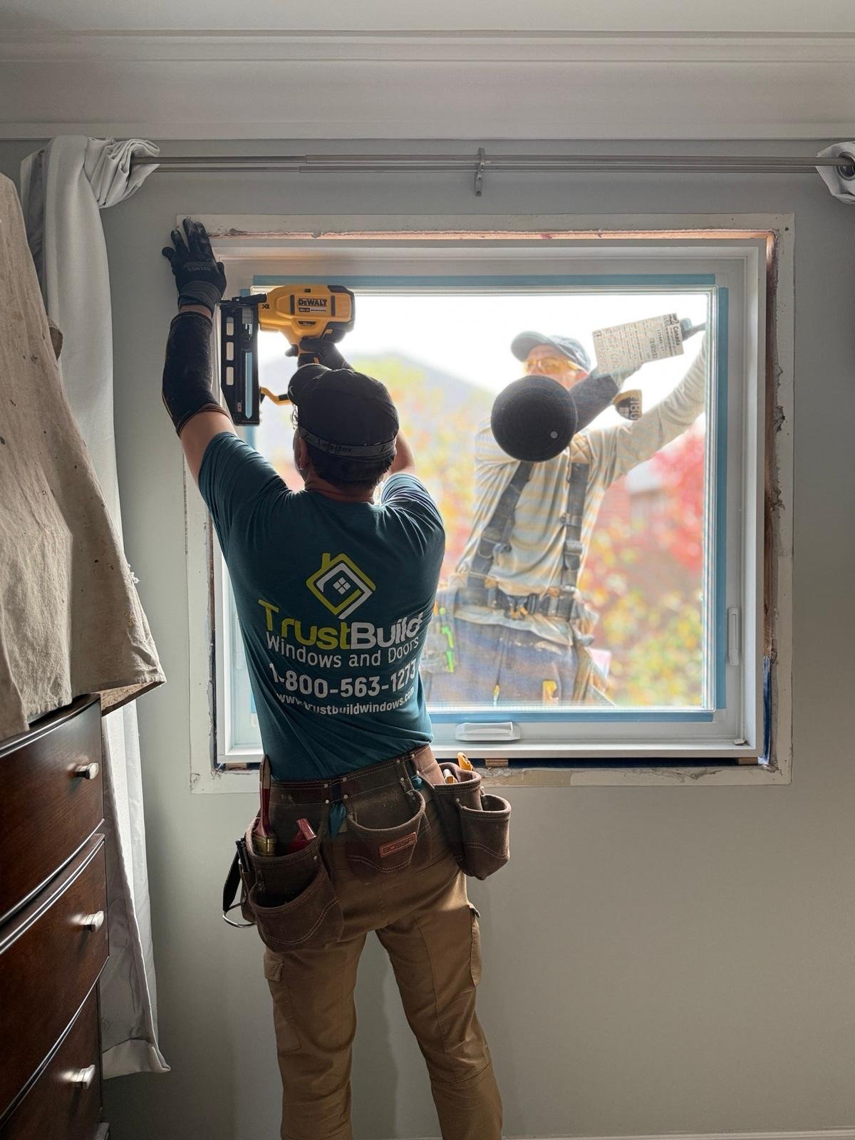 TrustBuild Windows and Doors contractors install a new window. One worker inside uses a nail gun on the frame, while another stands outside, viewed through the glass in a bedroom.