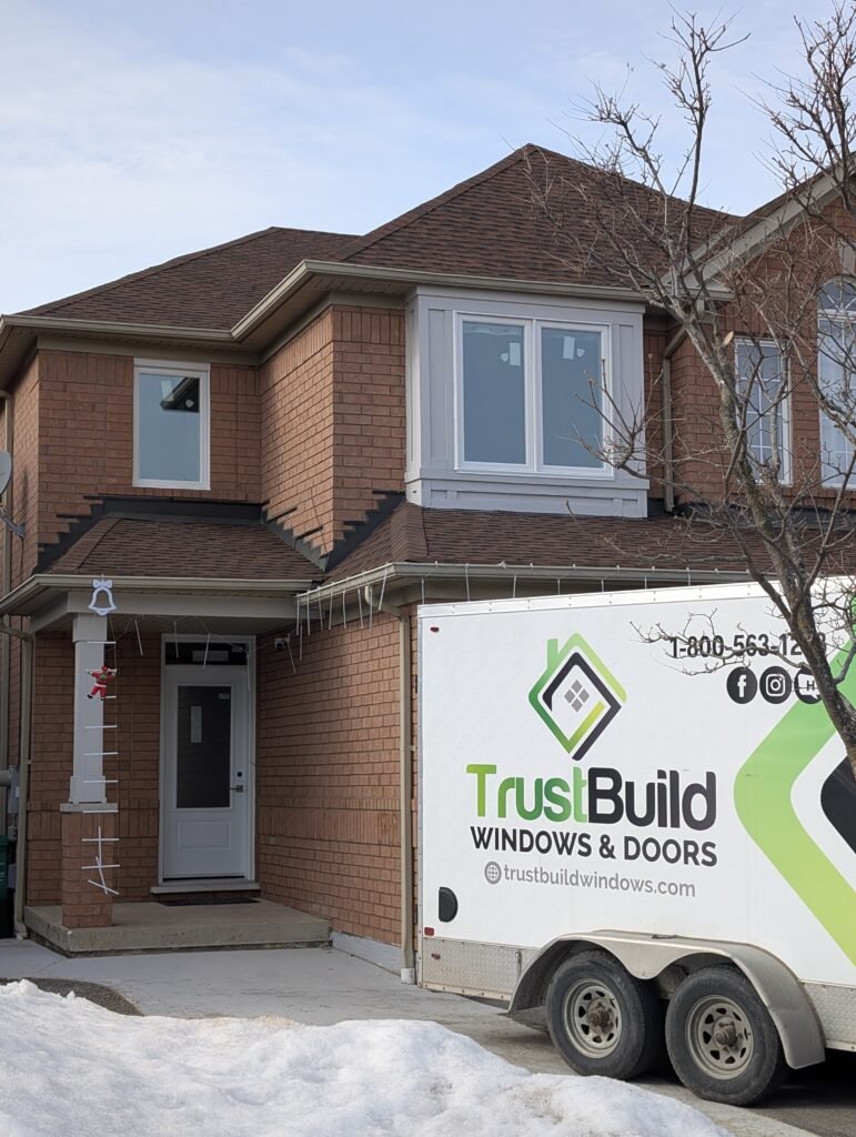 A TrustBuild Windows & Doors trailer is parked in front of a two-story brick house with newly installed white windows. Patches of snow and holiday decorations are visible on a winter day.