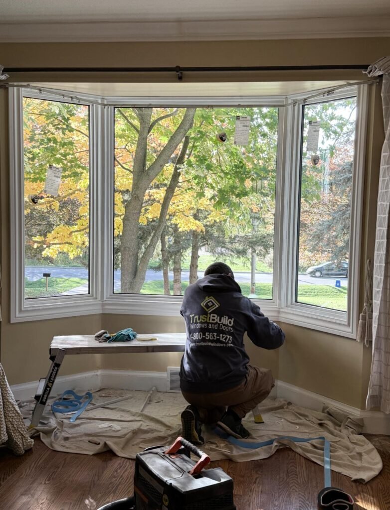 A TrustBuild Windows and Doors contractor kneels inside a home, installing a large white-framed bay window. Outside, a lush green tree and a residential street are visible through the glass.