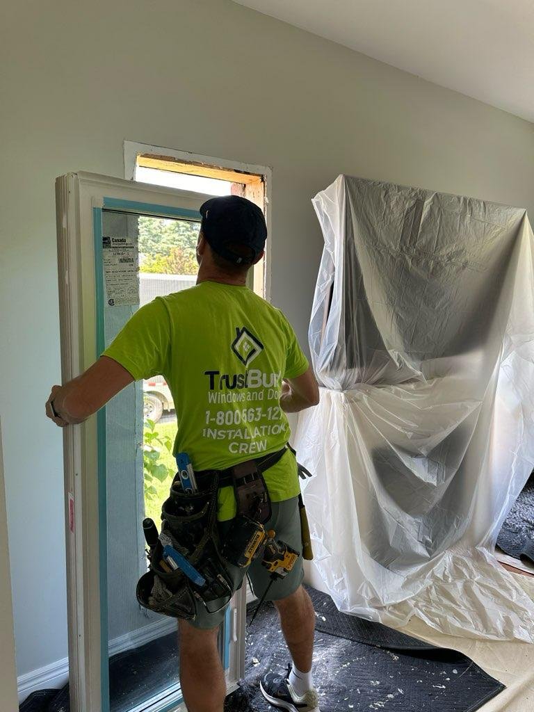 A TrustBuild Windows and Doors contractor in a bright lime green shirt fits a new white window frame into an opening while furniture remains protected under plastic sheets indoors.