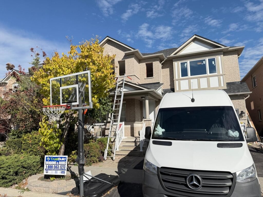 A white Mercedes-Benz work van is parked in front of a tan brick house during a window installation by TrustBuild. A tall ladder reaches the second story, and a company sign sits by the curb.