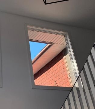 An interior view captures a rectangular window set high in a white wall above a staircase. The glass reveals a vibrant blue sky and the red brick side of a neighboring house under bright sunlight.