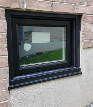 A sleek, black-framed hopper window is installed into a light brick and concrete foundation wall. The modern, dark finish provides a bold, contemporary contrast against the neutral masonry.