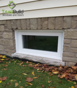 A white fixed-pane window is installed into a textured stone foundation wall. The clean vinyl frame provides a sharp contrast against the rough earth-toned masonry and autumn leaves.