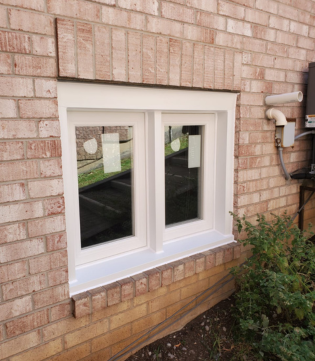 A white double-casement window is installed into a light-colored brick exterior wall. The new frame is thick and clean, reflecting the outdoor stairs and greenery in the clear glass panes.