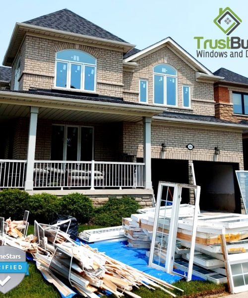 A large two-story stone house sits behind a pile of old window frames and debris on a blue tarp. New white-framed windows with arched tops are already installed on the home's second floor.