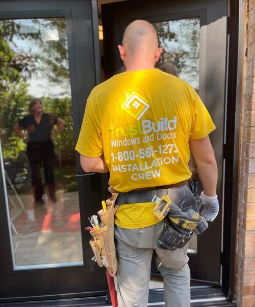 A worker in a yellow TrustBuild shirt stands with a tool belt, facing a new black-framed glass door installation. A client is visible through the glass reflecting the outdoor area.