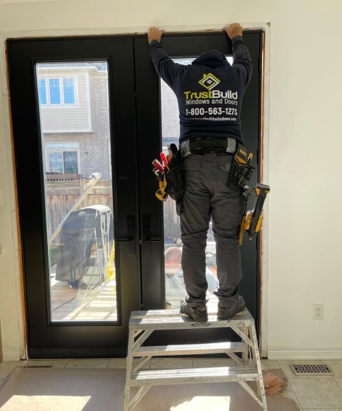 An installer in a navy TrustBuild hoodie stands on a stepladder to secure the top of a new black double-patio door. The interior installation overlooks a sunny backyard and deck.
