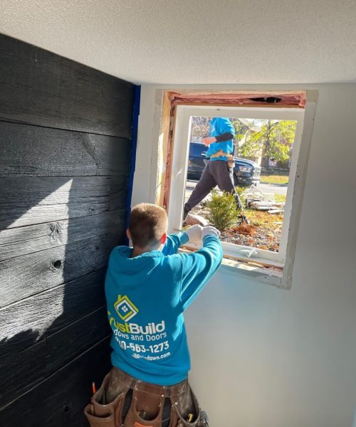 An interior view shows a window installer in a bright blue TrustBuild hoodie working on a white window frame. The open window reveals an outdoor scene with a second worker and a parked vehicle.