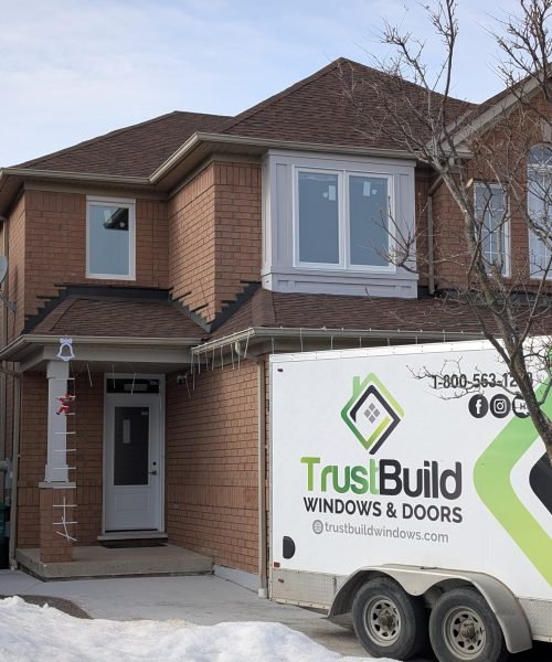 A TrustBuild Windows & Doors trailer is parked in front of a two-story brick house with newly installed white windows. Patches of snow and holiday decorations are visible on a winter day.