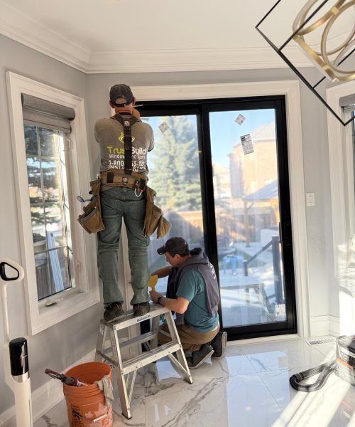 Two TrustBuild Windows and Doors contractors install a black sliding glass door. One worker stands on a step ladder to reach the top frame while the other kneels to secure the bottom track.