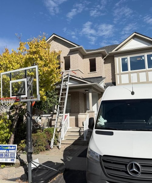 A white Mercedes-Benz work van is parked in front of a tan brick house during a window installation by TrustBuild. A tall ladder reaches the second story, and a company sign sits by the curb.
